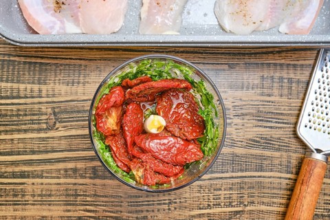 Fresh herbs, garlic and dried tomatoes are combined in a blender bowl. A baking tray with seasoned