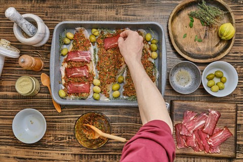 Hand places slices of bacon atop a seasoned fish fillets on a baking tray. Surrounding ingredients