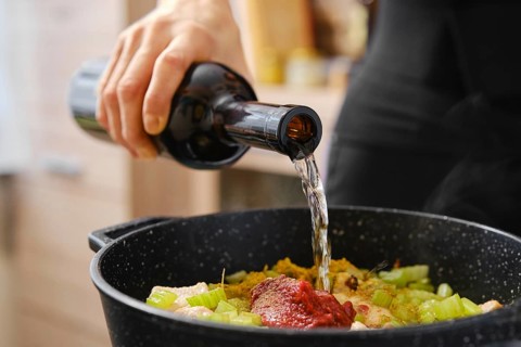 A person pours white wine from a dark bottle into a pot filled with chopped vegetables and