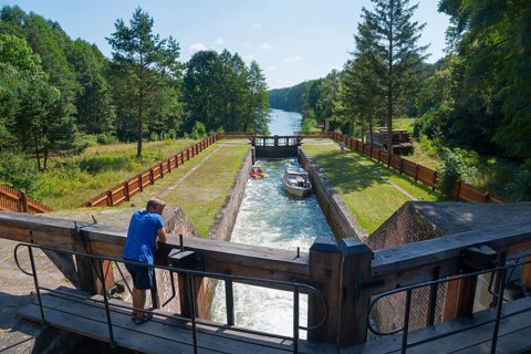 A man observes a lock with a boat on a river surrounded by trees, Sluza Paniewo, lock between the