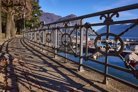 Forged railing with shadow, frog perspective, lakeside promenade, trees, lake, mountains, blue sky,