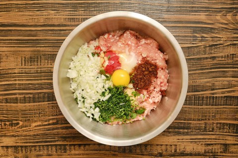 A metal mixing bowl holds a blend of ground meat, chopped onions, herbs, smoked paprika, egg, and