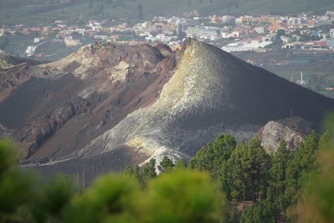 Steep volcano with visible lava and ash layers, city in the background, last volcanic eruption of