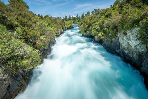 A rushing waterfall at Huka Falls surrounded by lush vegetation and blue sky, Taupo, New Zealand,