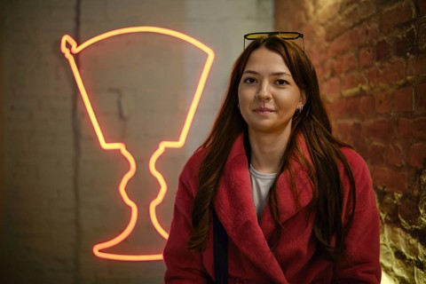 A young woman with long brown hair smiles while wearing a pink coat, seated in a trendy location