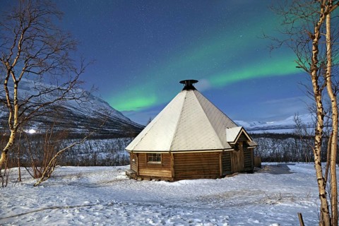 A snow-covered wooden hut under a glowing green aurora borealis in a wintry landscape with
