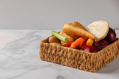 A woven basket full of vegetables such as soup vegetables, potatoes and onions on a marble tabletop
