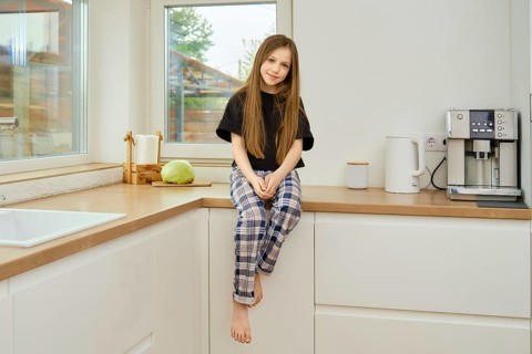 Cheerful young girl in plaid pants and black shirt sits on wooden kitchen tabletop
