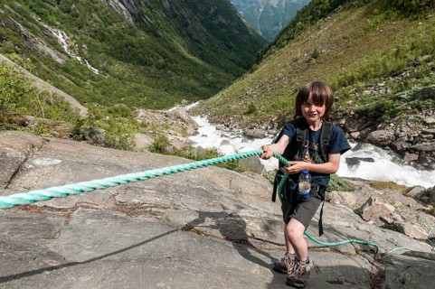 Boy uses rope at steep passage, rock, hiking path to glacier Buarbreen, river in valley stream down