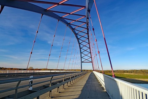 A modern steel bridge with pink and white struts spans a long distance under a clear sky, symbol of
