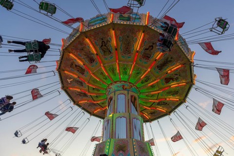 A colourfully illuminated carousel with swinging seats against an evening sky, wave flight, chain