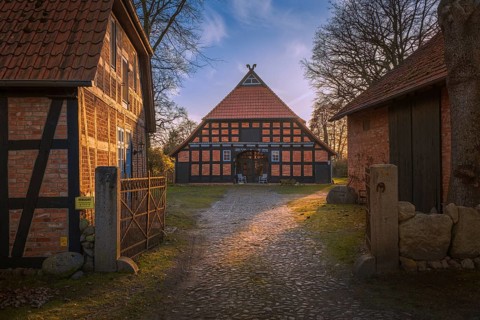 View of a courtyard entrance with a half-timbered farmhouse behind it in the evening light,