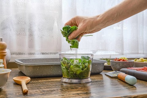 A hand gently places fresh basil leaves into a bowl for salad preparation. The kitchen features