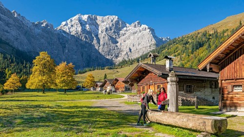 The alpine village of Eng in autumn, two hikers fill a water bottle at the wooden fountain,