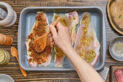 Fish fillets are arranged in a baking tray while a hand spreads a flavorful spice mixture over them