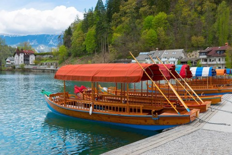 Wooden boat with red canopy on the lake in front of a wooded slope with houses, Lake Bled, Lake