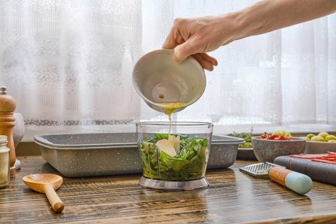 Hand pours lemon juice into a food processor filled with fresh green herbs and garlic. The kitchen