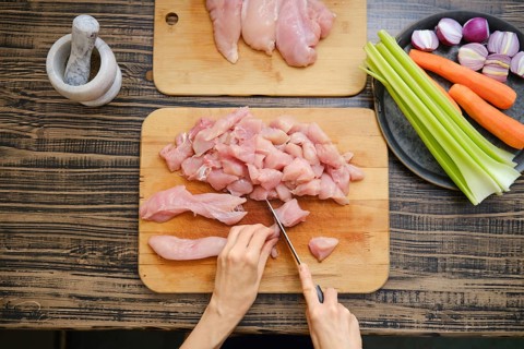 A person skillfully chops raw chicken on a wooden cutting board, surrounded by colorful vegetables