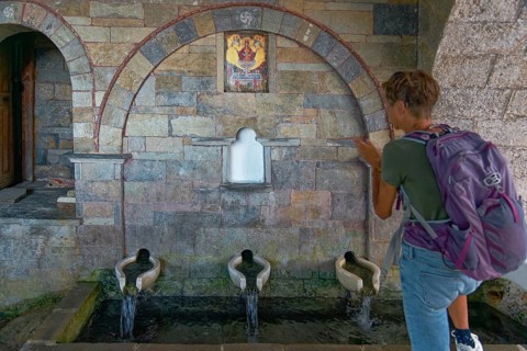 A woman stands in front of the old stone fountain with three water sources and marble basin below
