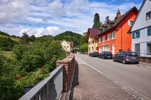 Apartment blocks, hard shoulder with parked cars, hilly landscape with forest, sandstone wall,