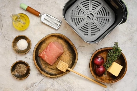 A raw pork loin on a wooden plate beside bowls filled with salt, pepper, and fresh thyme. An onion,