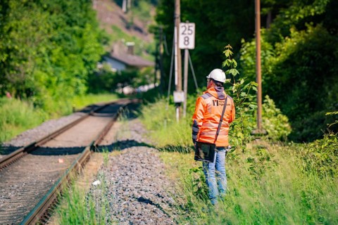 A worker in protective clothing and helmet stands next to railway tracks, surrounded by nature and