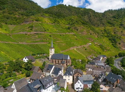 A small village with a church surrounded by vineyards and green hills under a blue sky with clouds,