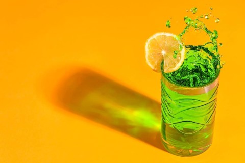 A green, sparkling cocktail with a slice of lemon casts shadows on an orange background