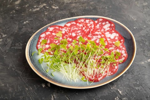 Slices of smoked cervelat salami sausage with spinach microgreen on black concrete background. Side