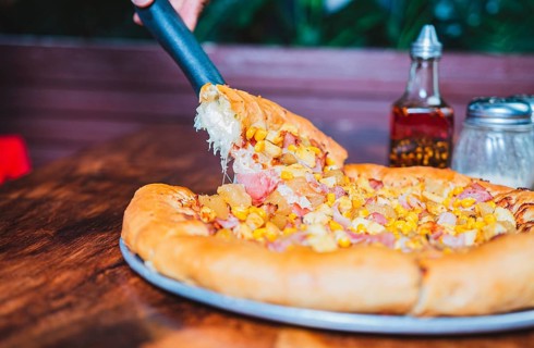 Person hand taking a slice of sweetcorn and ham pizza with spatula. Delicious ham pizza with cheese