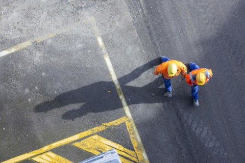 Two construction workers in hard hats walk over tarmac. They cast long shadows. Stuttgart,