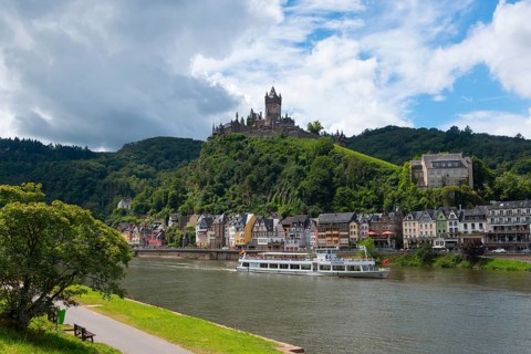 Lively town with a castle on a hill and ships on the river, surrounding green nature and clouds in