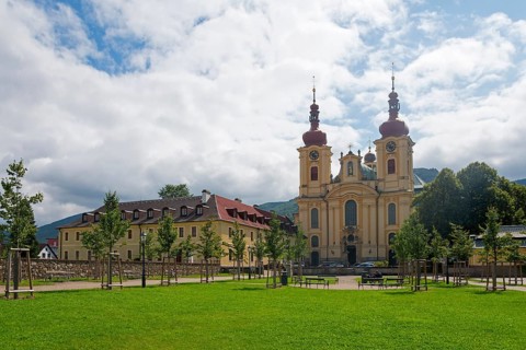 Baroque church with red roofs, surrounded by trees and green meadow with cloudy sky, monastery and