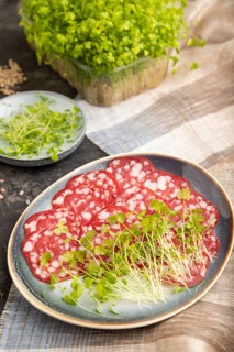 Slices of smoked cervelat salami sausage with spinach microgreen, salt and pepper on black concrete