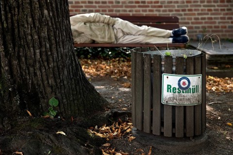 Homeless man lying on a bench in front of a rubbish bin labelled Restmüll, Alter cemetery, Bonn,