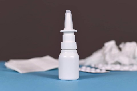 Nasal spray bottle with crumbled tissues and pills used against common cold in blurry background