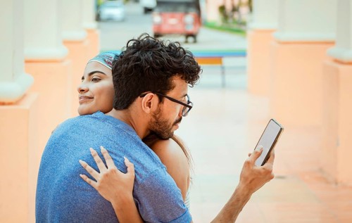 Unfaithful man looking at the cell phone while hugging his girlfriend outdoors. Unfaithful