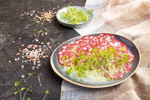 Slices of smoked cervelat salami sausage with spinach microgreen, salt and pepper on black concrete