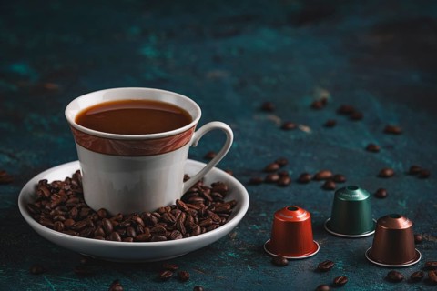 A cup of coffee next to three coffee capsules, surrounded by coffee beans on a textured surface