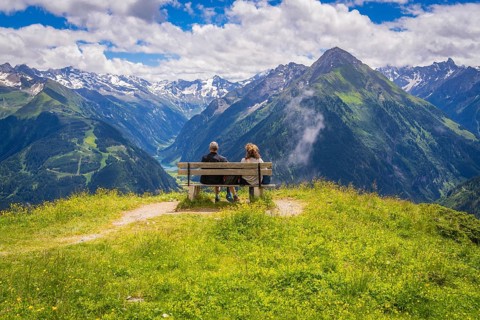 Two people sit on a bench and look out over a valley with majestic mountains in the background,