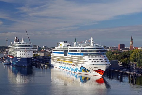 Two large cruise ships in the harbour, surrounded by water and city buildings in the background,