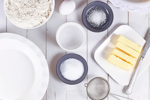 Pie crust ingredients in bowls. Flour, sugar, salt, ice water, butter and egg on wooden table