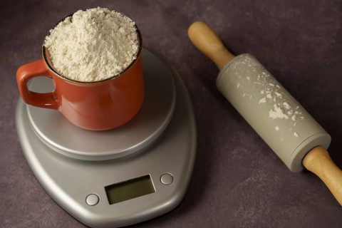 Red cup with flour on a kitchen scale next to a rolling pin with a dark background and copy space