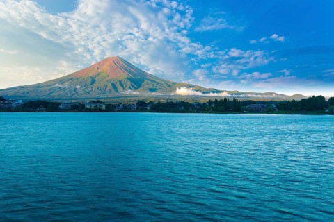 Mount Fuji bathes in morning side light while Lake Kawaguchiko sits in shadow below cloudy blue sky