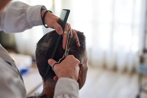 A hairdresser in a white shirt is cutting a man's hair with a pair of scissors and a comb at home