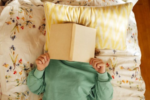 Caucasian blond teenage girl is lying on the bed and reading book. Reading fiction, hobbies,