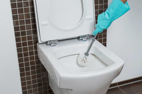 Person wearing a turquoise rubber glove cleaning under the rim of a toilet bowl with a toilet brush