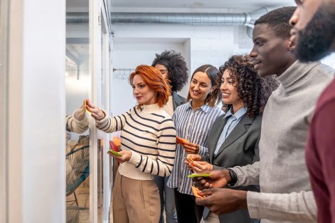Diverse coworkers using sticky notes on a glass board during brainstorming of a new project