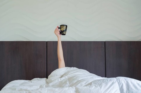 Drinking coffee early tomorrow. Woman in bed, under duvet, with arm raised up holding coffee cup