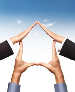 Conceptual home symbol made by hands in a shape of a house over blue sky and white background. Real